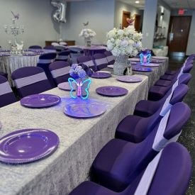 Long tables with purple plates and chairs, silver tablecloths, and butterfly centerpieces set for a party.
