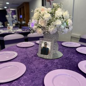 A round table with a floral centerpiece, a framed photo, and six lilac plates on a purple tablecloth.