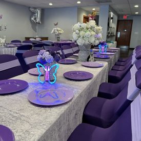 Table set for an event with purple decor, floral centerpieces, and butterfly-shaped neon lights.