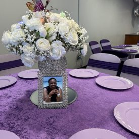 A table with lavender decor features a floral centerpiece, butterfly ornament, and framed photo on a mirror base.