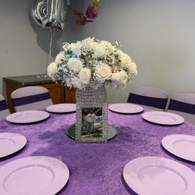Round table with a purple tablecloth, eight lavender plates, and a white floral centerpiece with a framed photo.