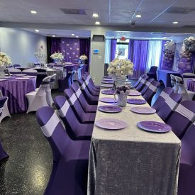 A banquet hall decorated in purple and silver with set tables, floral centerpieces, and balloon arrangements.