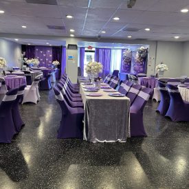 Event hall with purple and silver decor, round and rectangular tables, floral centerpieces, and balloon arrangements.