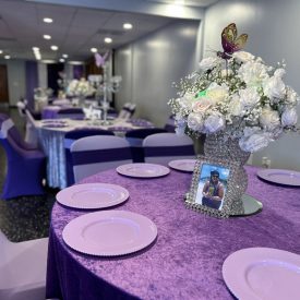 A round table with lavender plates and a floral centerpiece, set for an event in a decorated banquet hall.