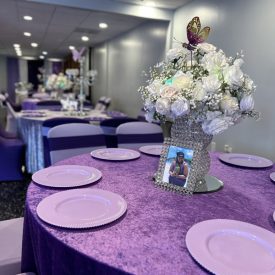 Round table with a purple cover, pink plates, floral centerpiece, and framed photo in a decorated banquet hall.