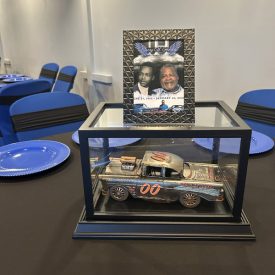 A model race car in a display case sits below a memorial photo on a table set with blue plates and chairs.
