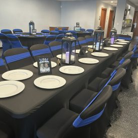 Rectangular table set with plates, lanterns, and framed photos; black chairs with blue sashes in a decorated room.