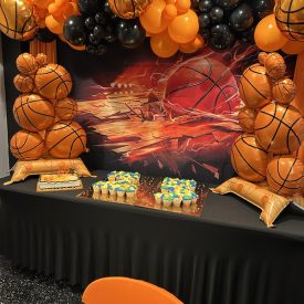 Basketball-themed party table with balloons, cupcakes, a cake, and basketball decorations against a sports backdrop.
