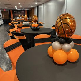 Tables with black and orange decor and basketball balloon centerpieces set up for an event in a banquet hall.